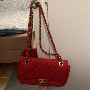 Röd quiltad axelväska med kedja - Snygg röd axelväska med quiltat mönster och guldfärgade detaljer. Väskan har en vridbar låsning framtill och en kedjerem med invävt rött material. Insidan har flera fack, inklusive ett med dragkedja. Perfekt för dig som vill sticka ut med en färgglad accessoar.