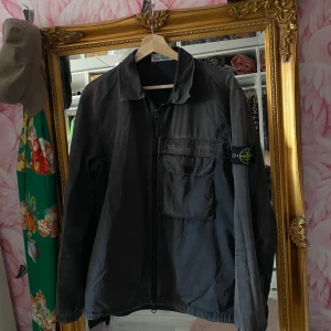Svart overshirt från Stone Island - Snygg svart overshirt från Stone Island med klassisk patch på ärmen. Jackan har dragkedja framtill, stor bröstficka med lock och knäppning samt en avslappnad krage. Perfekt lager-på-lager-plagg med clean look och streetkänsla.