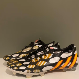 Adidas Preditor 2014 world cup  - Snygga Adidas Predator storlek 45 1/3 og world cup skor  fotbollsskor i svart med vita och gula hexagonmönster samt orangea ränder på sidan. Skorna har snörning och platt sula med dobbar för gräsplan. Tillverkade i syntetmaterial och designade för maximal kontroll på planen.