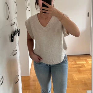 Beige stickad väst med v-ringning - Säljer en beige stickad väst med breda ärmöppningar och v-ringad hals. Västen har ribbade kanter vid hals och nederkant, och är perfekt att styla över en skjorta eller t-shirt. Enkel och stilren design som funkar året runt. 