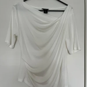Lindex vit topp, storlek S - Vit topp med drapering framtill och kort ärm från Lindex i storlek S. Elegant och stilren design som passar både till vardag och fest. 95% viskos och 5% elastan.  Toppen är i nyskick, oanvänd.   Mätt: Längd ca 60 cm Byst ca 46 cm Midja ca 40 cm Ärmlängd mätt från axeln ca 24 cm Axel till axel ca 50 cm