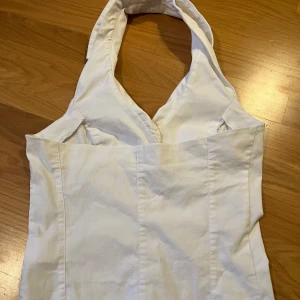 Vit halterneck topp med knappar - Snygg vit topp med halterneck och djup v-ringning. Den har fyra vita knappar framtill, två dekorativa fickor och markerad krage. Figurnära passform och sydda sömmar för en clean look. Hör av dig vid frågor, lägg prisförslag ❤️