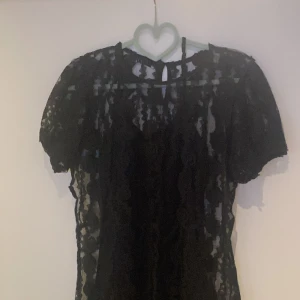 Svart spets klänning med kort ärm - Supersnygg svart klänning  i genombruten spets med korta ärmar och rund hals. Blusen har ett romantiskt mönster och är perfekt för dig som vill ha en cool och elegant look. Lätt transparent och med fina detaljer i spetsen. Underklänning medföljer 