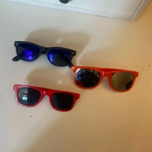 3 par färgglada solglasögon - Tre par solglasögon i plast med klassisk wayfarer-form. Ett par är svarta med blåtonade spegelglas, ett par är röda med svarttonade glas och texten 'JESUS ÄR COOL' på skalmen, och ett par är orange med spegelglas och texten 'liftroller'. Perfekta för sommaren!