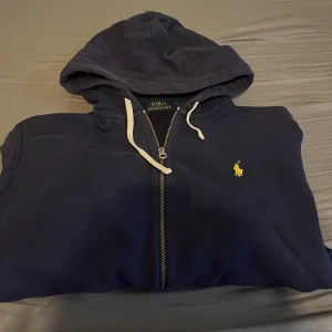 Marinblå hoodie från Polo Ralph Lauren - Säljer en marinblå hoodie från Polo Ralph Lauren med dragkedja och vit snörning i huvan. Klassisk gul logga broderad på bröstet och stora fickor framtill. Perfekt för dig som gillar stilren streetwear och vill ha något bekvämt och snyggt. Hör av er vid minsta lilla fråga 