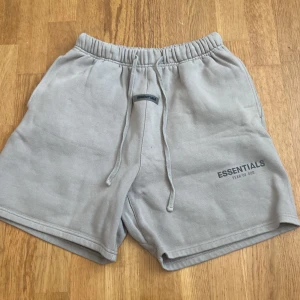 Essentials shorts Grå/Beige - Snygga grå/beige shorts från Essentials Fear of God i mjuk bomullsblandning i väldigt bra skick. De har elastisk midja med snörning och logga framtill samt på benet. 