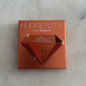 Huda Beauty Topaz Obsessions palett - Säljer en Huda Beauty Topaz Obsessions ögonskuggspalett med nio nyanser i varma toner som orange, koppar, guld, brons och brunt. Paletten innehåller både matta och skimrande skuggor i ett kompakt format med spegel i locket.
