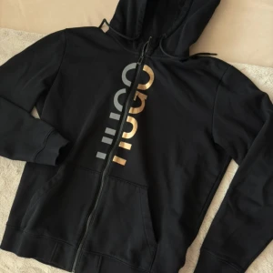 Svart hoodie från Hugo med guldtryck - Svart hoodie från Hugo med stort vertikalt logotryck i guld och grått på framsidan. Tröjan har huva med dragsko, hel dragkedja och två fickor framtill. Tillverkad i mjuk bomullsmix, perfekt för en chill och stilren look.