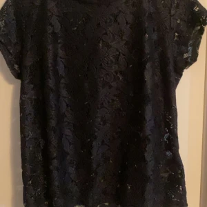 Svart spetsblus med dragkedja - Säljer en svart blus i spets med korta ärmar och snygg dragkedja i ryggen. Blusen har ett blommigt spetsmönster som ger en elegant vibe och är perfekt om du vill sticka ut lite extra. Passformen är normal och materialet känns lätt och luftigt.