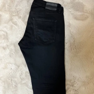 Svarta slim jeans Jack & Jones - Svarta jeans från Jack & Jones i modellen Slim/Glenn. Snygg, smal passform med klassisk femficksdesign och diskreta nitar. Jeansen är tillverkade i stretchigt bomullsmaterial för extra komfort. Perfekta för dig som gillar en stilren look.