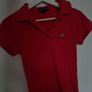 Röd pikétröja från Lyle & Scott - Snygg röd pikétröja från Lyle & Scott med klassisk krage och korta ärmar. Tröjan har det ikoniska gula fågel-loggan broderad på bröstet och är gjord i bomull för en skön känsla. Perfekt för en stilren och sportig look. Äkta, Orginalpris 749 kr. ❤️