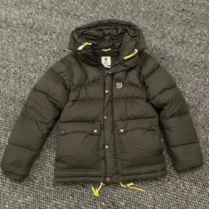 Grön dunjacka från Fjällräven XS - Säljer en grön dunjacka från Fjällräven i storlek XS man men sitter som s dam. Jackan har huva, gula snören och klassisk logga på bröstet. Flera fickor framtill med tryckknappar och dragkedja. Perfekt för kalla dagar och riktigt snygg passform. Som ny, endast använd typ två månader 
