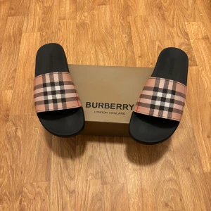Burberry sandaler med klassiskt rutmönster - Snygga sandaler från Burberry med svart sula och bred rem i det ikoniska beige, svarta och röda rutmönstret. Perfekta för sommaren med en stilren och lyxig känsla. Remmen är i syntetmaterial och sulan är platt för maximal komfort.