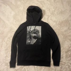 Svart Tupac hoodie  - Svart hoodie med stor Tupac-tryck på bröstet. Klassisk känguruficka framtill, snörning i huvan och långa ärmar. Hoodien har en relaxed vibe och är perfekt för dig som gillar streetwear och hiphopstil.