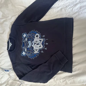 Mörkblå Kenzo sweatshirt med tiger - Säljer en mörkblå sweatshirt från Kenzo med ikoniskt broderat tigerhuvud i blå och vita toner på bröstet. Tröjan har rund hals, långa ärmar och ribbade muddar. Perfekt för dig som gillar streetstyle och vill sticka ut med ett snyggt statement-plagg. Liten fläck som jag inte testat ta bort. 