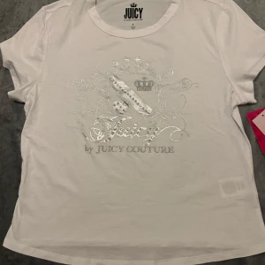 Vit Juicy Couture t-shirt med silvertryck - Vit t-shirt från Juicy Couture med glittrigt silvertryck och emblem framtill. Klassisk rund halsringning och korta ärmar. Snygga detaljer med pärlliknande dekorationer och logga. Perfekt för dig som gillar en clean men ändå lyxig look. Är L men passar som s och xs.