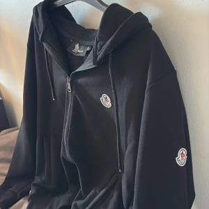Svart Moncler hoodie med dragkedja - (STORLEK: S) passar någon som är runt 170-175. Svart hoodie från Moncler med klassisk logga på bröstet och ärmen. Tröjan har huva med snörning, hel dragkedja framtill och två fickor. Tillverkad i mjuk bomull 