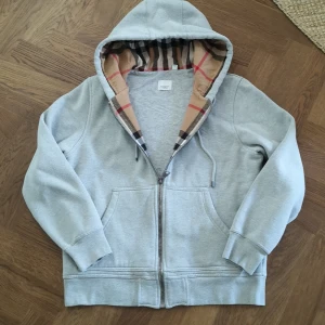 Burberry hoodie - Snygg grå hoodie från Burberry med klassiskt rutigt foder i huvan och broderad logga på bröstet. Tröjan har dragkedja framtill, snörning i huvan och två fickor. Perfekt för dig som vill ha en stilren och lyxig look.