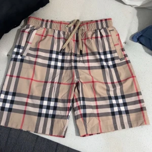 Rutiga burberry shorts - Snygga shorts från Burberry i klassiskt beige rutigt mönster med inslag av svart, vitt och rött. De har elastisk midja med snörning och sidofickor samt en bakficka. Perfekta för sommaren och ger en clean och trendig vibe. Liten i storleken. Passar om en L/Xl