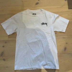 Vit Stüssy t-shirt med tärningsprint - Vit t-shirt från Stüssy med svart logga på bröstet och stor grafisk print med röda tärningar och logga på ryggen. Klassisk passform och rund hals. Perfekt för dig som gillar streetwear och vill sticka ut med en cool design.