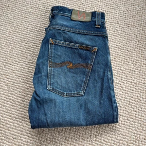 Blå jeans från Nudie Jeans - Säljer ett par klassiska blå jeans från. Modellen har raka ben och ikonisk bakficka med vågig söm. Perfekta för en avslappnad och stilren look. En olagad ficka