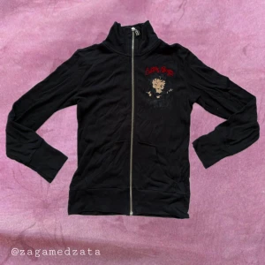 Svart Betty Boop zipjacka från o'she - Svart långärmad zipjacka med stort Betty Boop-tryck och röda broderade detaljer fram och bak. Jackan har hög krage, dragkedja med BB-berlock och är gjord i mjuk bomull. Perfekt för dig som vill sticka ut med en ikonisk och lekfull look. Passar mer en storlek S-M.