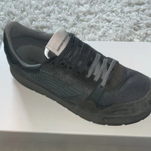 Mörkgröna sneakers från Emporio Armani - Snygga svarta sneakers från Emporio Armani med grå detaljer. Skorna har en mix av mocka och mesh, samt klassisk snörning. Ikoniska Armani-loggan syns på sidan och tungan. Perfekta för dig som gillar stilrena och exklusiva sneakers.