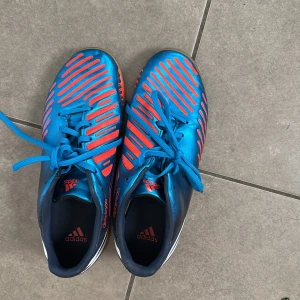 Blå och röda adidas fotbollsskor, storlek 37,5 - Snygga adidas fotbollsskor i blått och rött med klassiska vita ränder på sidan. Skorna har snörning, platt sula i gummi och coola röda detaljer på ovandelen. Perfekta för inomhusspel och har en sportig vibe som sticker ut på planen.