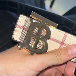 Burberry bälte med rutigt mönster - Säljer ett Burberry bälte med ikoniskt rutigt mönster. Ena sidan är beige med röda och svarta rutor, andra sidan är svart med diskret rutmönster. Snyggt guldfärgat spänne med Burberrys logotyp. Perfekt accessoar för att lyfta din outfit.