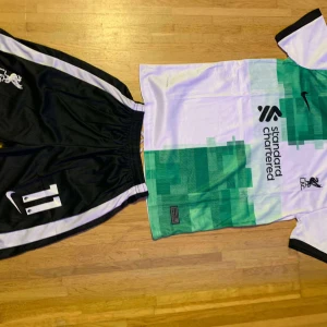 Liverpool bortatröja Nike nr 11 - Liverpool FC bortatröja från Nike med digitalt grönt och vitt mönster, svarta detaljer och tryckta loggor. Nummer 11 på ryggen och byxorna. Tröjan är kortärmad och tillverkad i lätt, ventilerande material perfekt för fotbollsträning eller match.