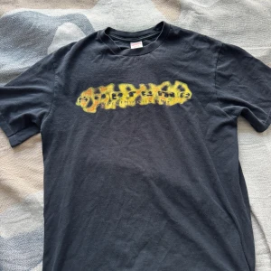 Supreme svart t-shirt med flames-tryck - Svart t-shirt från Supreme med korta ärmar och rund hals. På bröstet finns ett fett flames-tryck i gult och orange som ger en riktigt street vibe. Skön bomullskvalitet och relaxed fit, perfekt till jeans eller shorts.