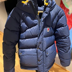 Blå dunjacka från Fjällräven med huva - Snygg blå dunjacka från Fjällräven med huva och gula snören. Jackan har två stora fickor framtill, dragkedja och tryckknappar. Klassisk Fjällräven-logga på bröstet och quiltad design som håller dig varm under kalla dagar.