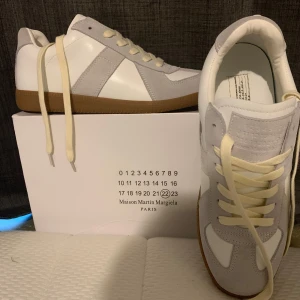 Maison Margiela vita och grå sneakers - Maison Margiela sneakers i vitt och ljusgrått skinn och mocka, med klassisk gummisula i brunt. Snygg retrostil med rund tå och krämvita snören. Perfekta för dig som gillar minimalistisk och stilren design.