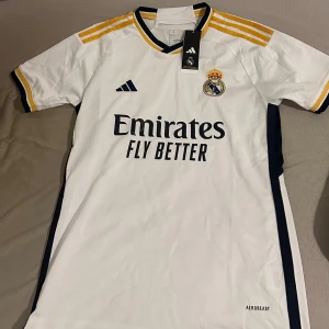 Real Madrid hemmatröja Adidas vit - Snygg vit Real Madrid hemmatröja från Adidas med gula och svarta detaljer. Klassisk design med klubbmärke på bröstet och 'Emirates Fly Better' tryck. Tillverkad i lätt och ventilerande material, perfekt för fotboll eller supporterliv.