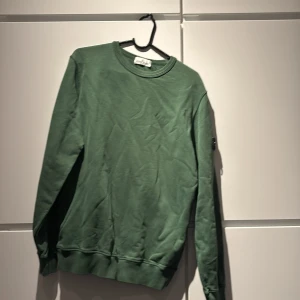 Grön Stone Island sweatshirt - Snygg grön sweatshirt från Stone Island Junior med klassisk rund hals och ribbade muddar. Tröjan har den ikoniska Stone Island-loggan på vänster ärm. 