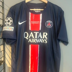 PSG Final Munich 2025 matchtröja - Paris Saint-Germain fotbollströja från Final Munich 2025 med D. Doué och nummer 14 på ryggen. Mörkblå bas med bred röd rand och vita detaljer, officiella loggor och Qatar Airways sponsortryck. Kortärmad modell i lätt funktionsmaterial, perfekt för fotbollsälskare. Helt ny med tagg en replika 
