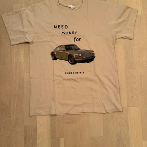 Beige t-shirt med Porsche 911-tryck - Beige t-shirt från EVOCUST med svart text 'NEED MONEY FOR PORSCHE 911' och en stor bild på en klassisk Porsche 911 på framsidan. Klassisk passform med rund hals och korta ärmar. Tillverkad i mjuk bomull, perfekt för dig som gillar bilar och streetstyle.
