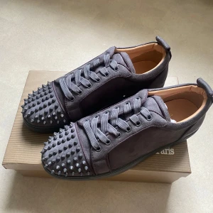Gråa Christian Louboutin sneakers med nitar - Snygga grå sneakers från Christian Louboutin i mocka med ikoniska röda sulor och coola nitar över tån. Skorna har snörning och en platt sula, perfekt för dig som vill sticka ut med en edgy och lyxig stil.