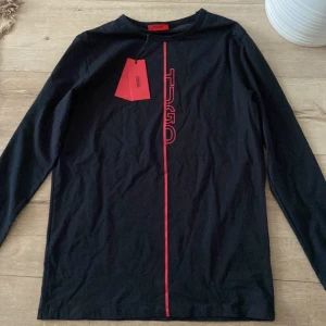 Svart långärmad tröja från Hugo Boss - Svart långärmad tröja från Hugo Boss med röd vertikal linje och HUGO-logga framtill. Tröjan är i mjuk bomull och har en clean, stilren look med normal passform. Perfekt för dig som gillar minimalistisk design med en touch av färg.