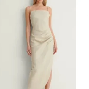 Beige långklänning i linne från NA-KD - Snygg långklänning från NA-KD i beiget linnematerial. Klänningen har smala axelband, rak urringning, draperad detalj på sidan och slits framtill. Den har även en öppenrygg. Perfekt för sommarens alla tillfällen. Knytning i ryggen för extra passform. Storleken är 38 men funkar även på 36. Originalpris 900kr.