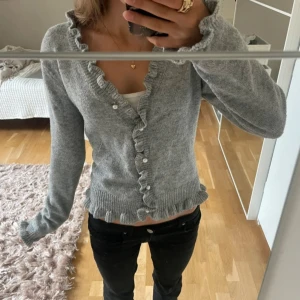 Grå kofta med volangdetaljer - Supersöt grå kofta med volangkant längs knäppningen och nederkanten. Långärmad och ribbad vid ärmslut och midja. Perfekt att slänga över ett linne.