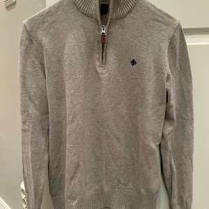 Beige stickad half zip från Morris - Snygg beige stickad tröja från Morris med ribbad krage och half zip-dragkedja med läderdetalj. Liten svart broderad logga på bröstet. Perfekt för lager-på-lager och chill stil. Långärmad och i mjukt material.