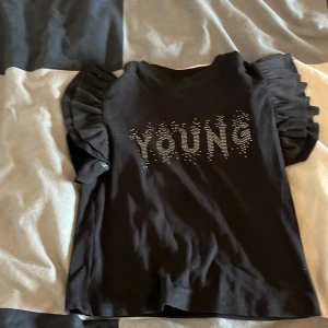 Svart croppad topp med volangärmar - Snygg svart croppad topp från Gina Tricot med glittrande texten 'YOUNG' framtill. Toppen har korta volangärmar som ger en cool och trendig look. Perfekt för dig som vill sticka ut lite extra med detaljer och glitter.