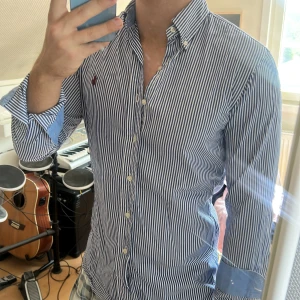Randig skjorta från Ralph Lauren - Säljer en klassisk blåvit randig skjorta från Ralph Lauren med broderad logga på bröstet. Skjortan har lång ärm, button-down krage och vita knappar. Tillverkad i bomull och har ljusblå detaljer på insidan av manschetterna. Perfekt för en clean och stilren look.