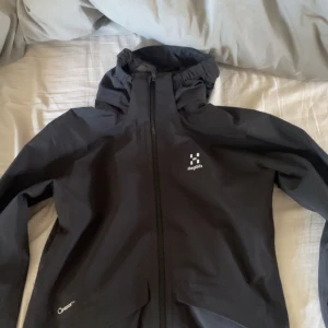 Svart vindjacka Haglöfs  - Svart vindjacka från Haglöfs i storlek 158 för junior. Jackan har huva, hel dragkedja och meshfoder på insidan. Tillverkad i slitstark polyester med Proof™-teknologi som skyddar mot vind och regn. Perfekt för uteliv och äventyr.