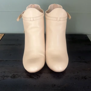 Beige stövletter med klack från Bonprix - Snygga beige stövletter från Bonprix i storlek 38. Skorna har rund tå, hög klack och dragkedja baktill. Snygga cut-outs på sidorna och dekorativ knytning ger en modern look. Tillverkade i syntetmaterial med stilren finish. Använda en gång.