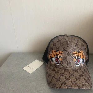 Gucci keps med tigertryck och GG-mönster - Snygg keps från Gucci med klassiskt GG-monogram i brunt och två coola tigertryck framtill. Baksidan är i svart mesh med justerbart band i grönt och rött. Kepsen har märkets logga invändigt och är tillverkad i polyester och bomull. PRIS KAN DISKUTERAS!