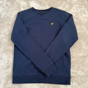 Mörkblå sweatshirt från Lyle & Scott - En stilren mörkblå sweatshirt från Lyle & Scott med klassisk rund hals och gul logga på bröstet. Tröjan har långa ärmar och är gjord i mjuk bomull, perfekt för en chill och avslappnad look.