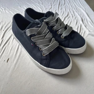 Marinblå sneakers Tommy Hilfiger - Snygga marinblå sneakers från Tommy Hilfiger med breda, randiga snören i vitt och svart. Skorna har guldfärgade öljetter, vit sula och diskret logga på sidan. Ovandel i textil och sula med TOMMY-print undertill. Perfekta för en stilren och sportig look.