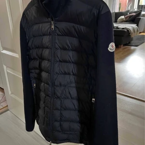 Moncler cardigan naivy  - Svart dunjacka från Moncler med quiltad framsida och stickade ärmar. Jackan har hög krage, dragkedja framtill och Moncler-logga på ärmen. Perfekt för dig som vill ha en clean och stilren look med premiumkänsla.